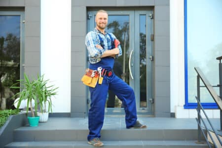 A smiling handyman in coveralls and a toolbelt stands confidently in front of a building with a tall glass door.