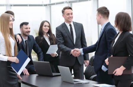 Young professionals smile as two men in suits shake hands over a black desk with open laptops on it.