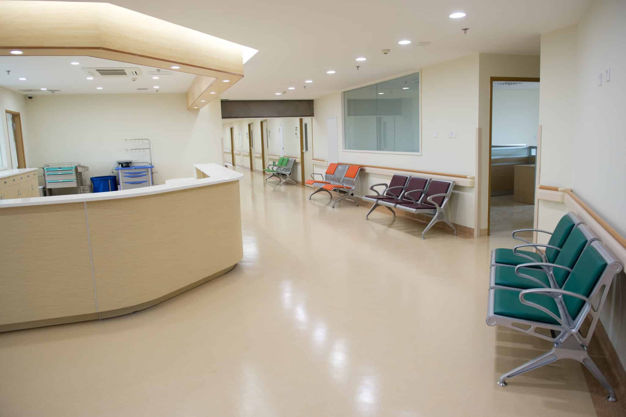 A brightly lit medical-clinic corridor with tan linoleum, nurses’ desk, and seating along the wall.